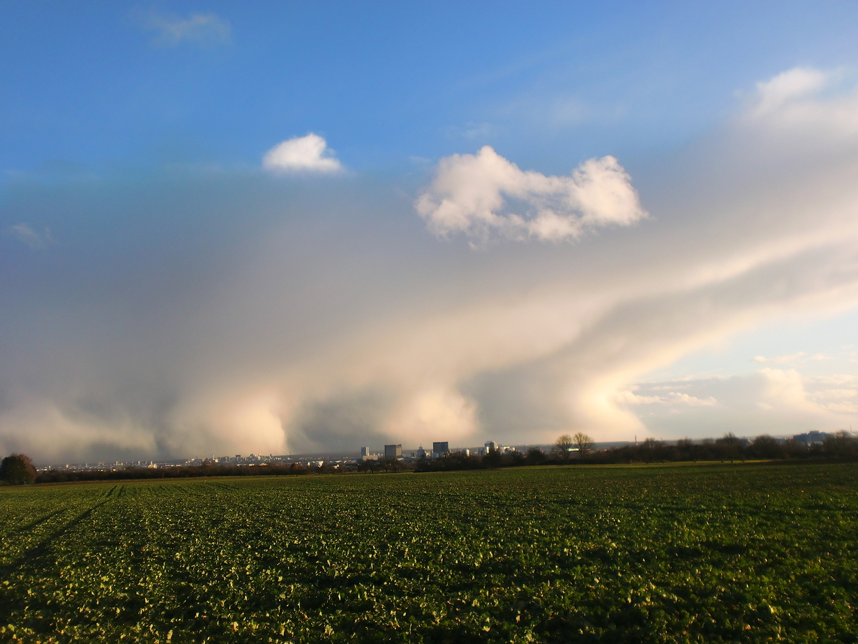 Grünes Feld mit Stadt im Hintergrund und dramatischen Wolkenformationen am Himmel.