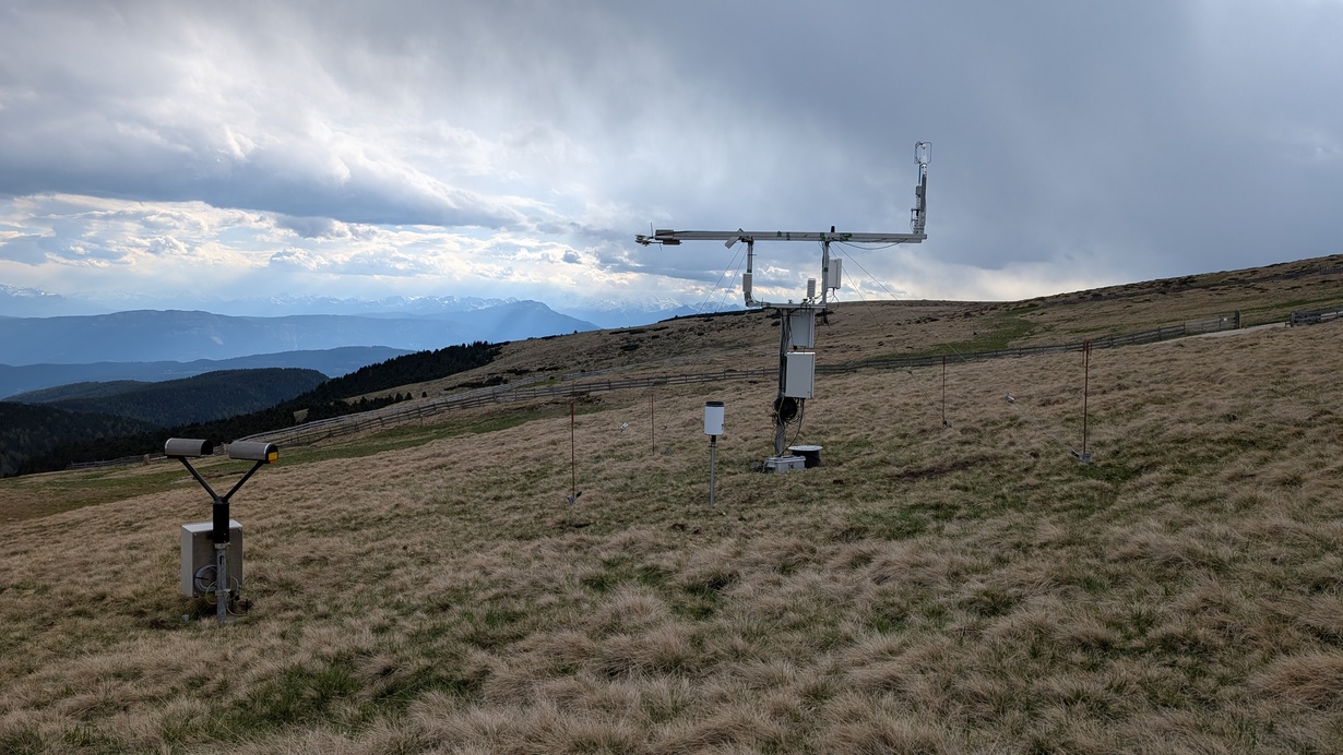 Wetterstation mit Messinstrumenten auf einer Bergwiese unter bewölktem Himmel.